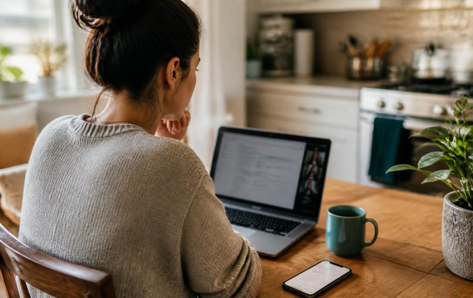A remote team member reading the meeting recap on a laptop at a kitchen table, with follow-ups open on the phone beside them